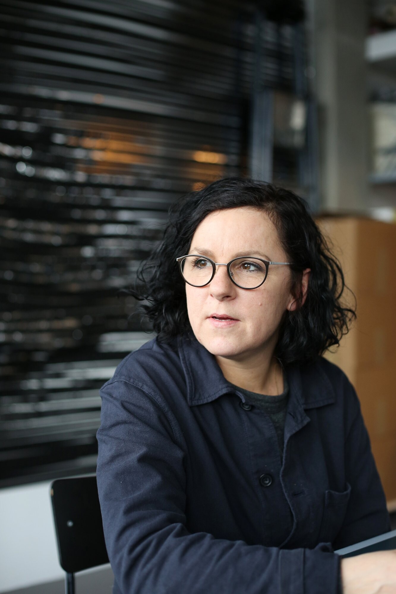 Portrait of a person with curly dark hair and glasses looking to the side, with a dark glossy wall structure behind them.