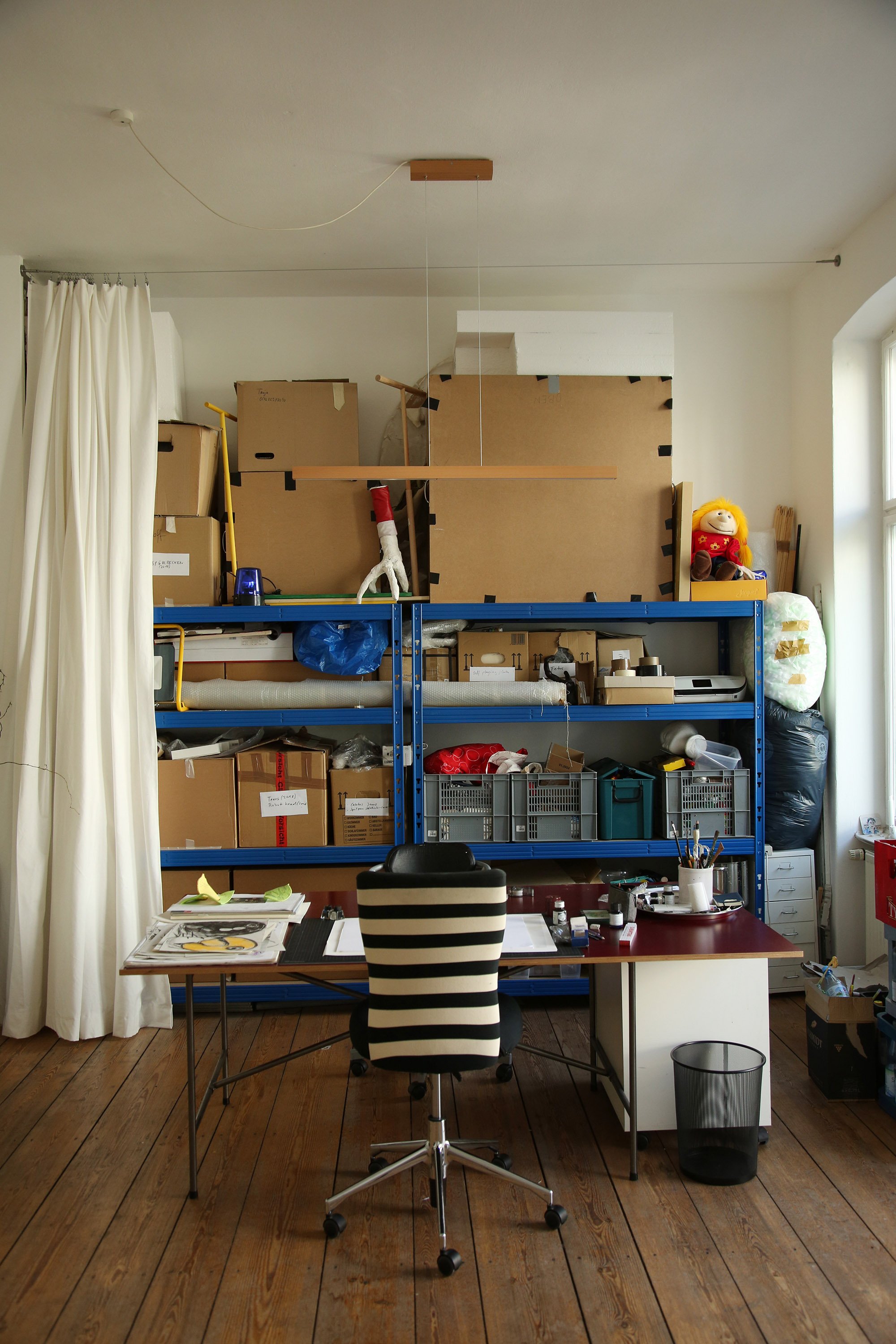 Workspace with a striped office chair at a desk, with a large blue shelving unit behind it filled with boxes, tools, and materials.