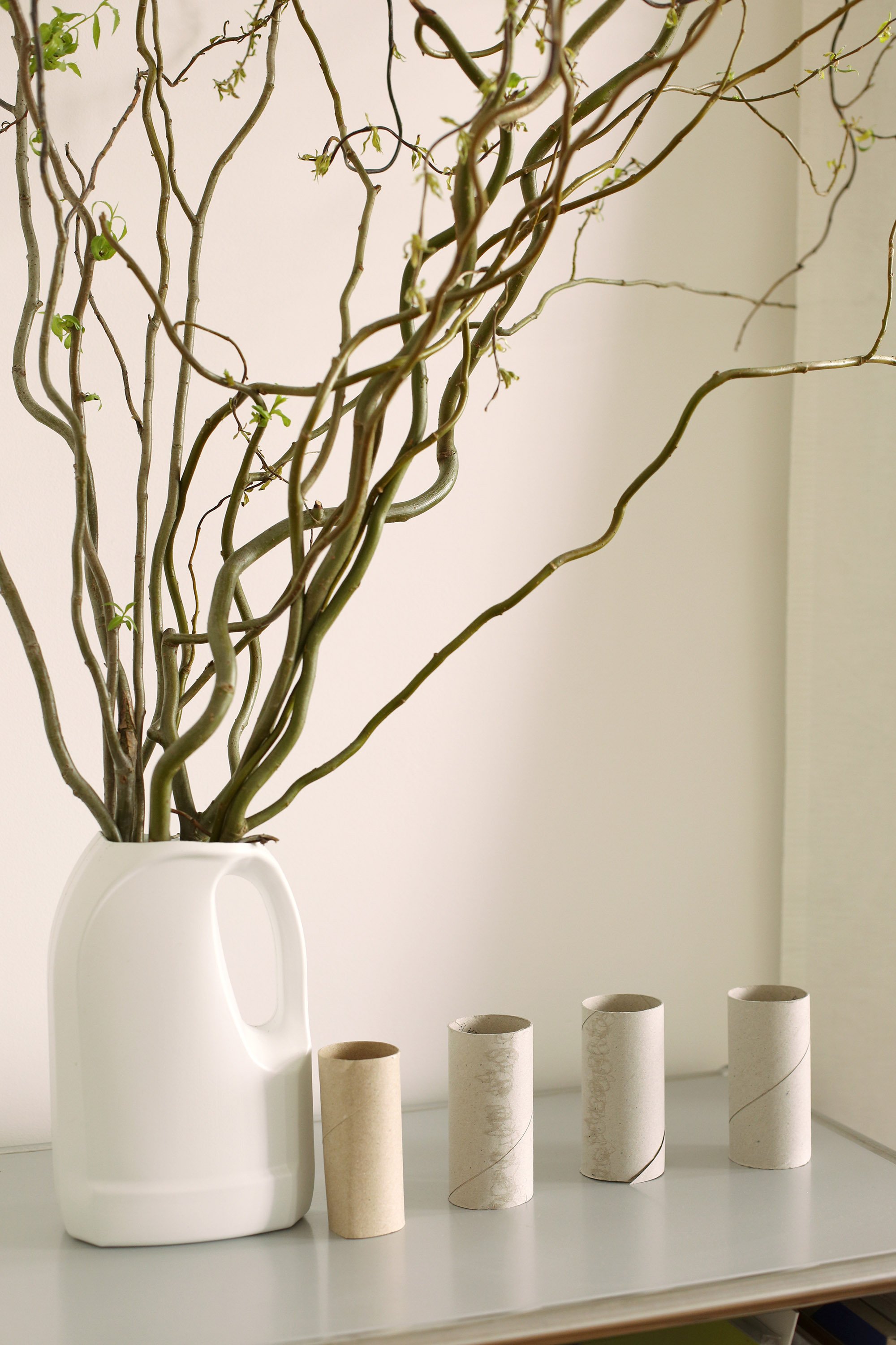 A white jug filled with branching twigs stands next to four empty cardboard tubes arranged in a row.