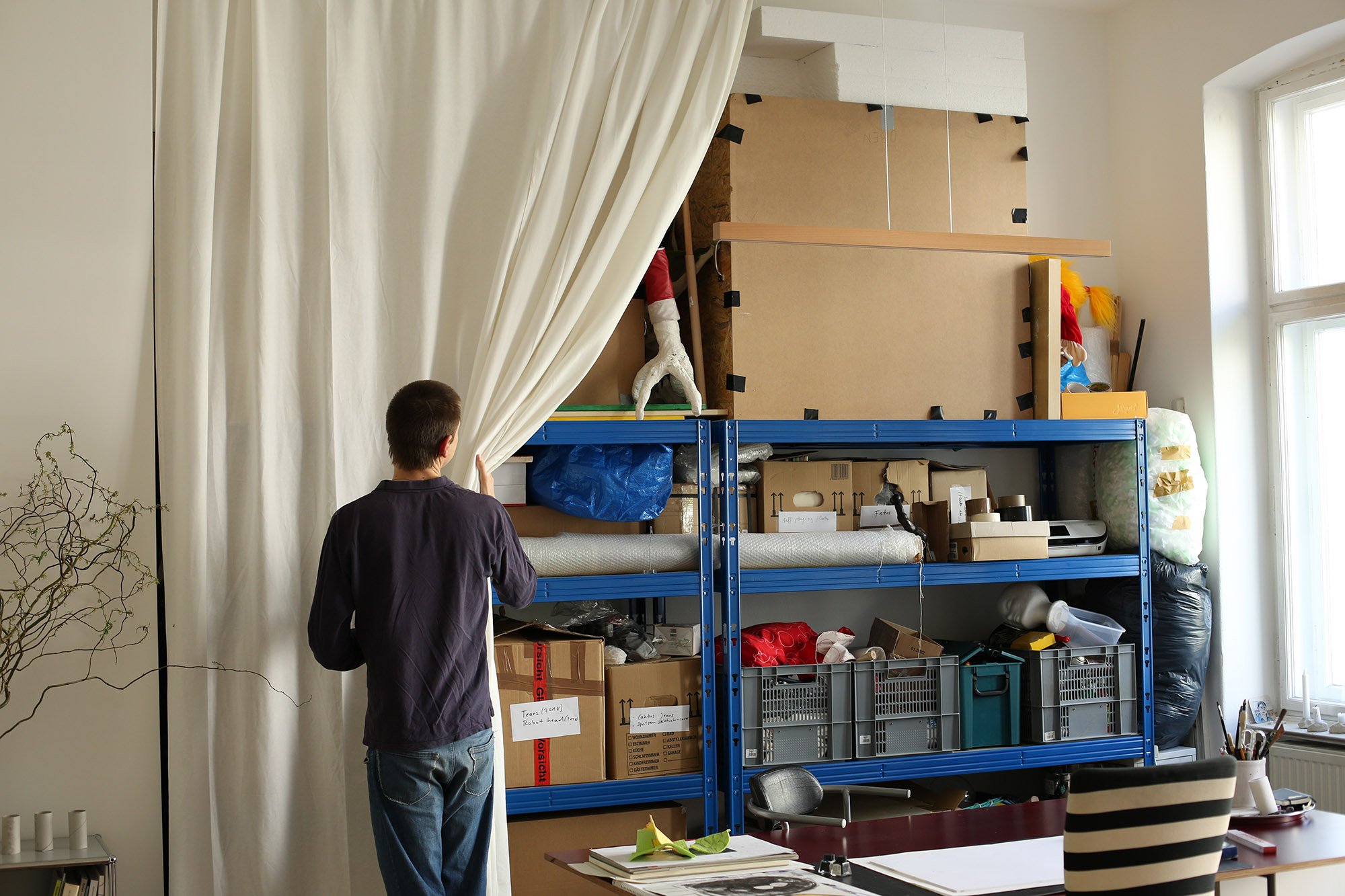 A person pulls aside a white curtain, revealing a large blue metal shelf filled with boxes and materials.