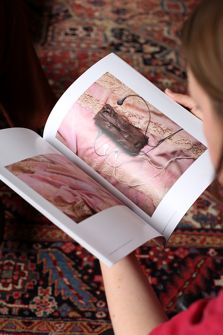 Someone holds an open book featuring a photograph of a tied object and earbuds resting on pink fabric. The patterned carpet is visible in the background.