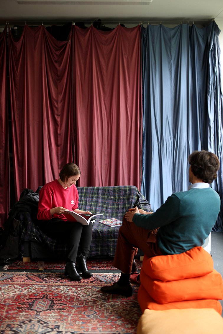 Two people sit facing each other. One person sits on orange cushions, the other on a sofa with a checkered cover, reading a book. Large red and blue fabric panels hang behind them.