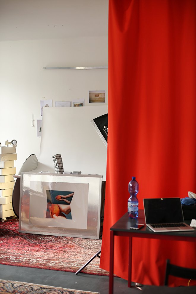 A studio corner with a red curtain, a black table holding a laptop and a water bottle. Behind it are photo prints and a light reflector against the wall.