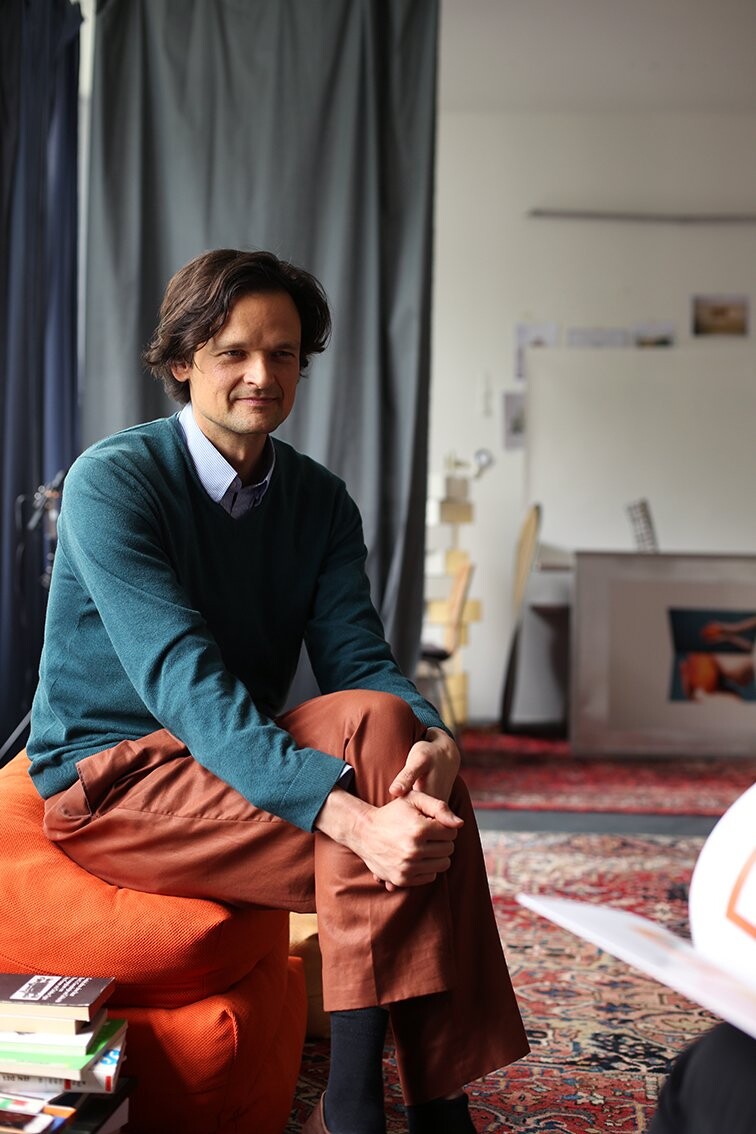 A person wearing brown pants and a green sweater sits on stacked orange floor cushions, with books placed on the floor beside them. A patterned carpet and gray curtains are visible in the background.
