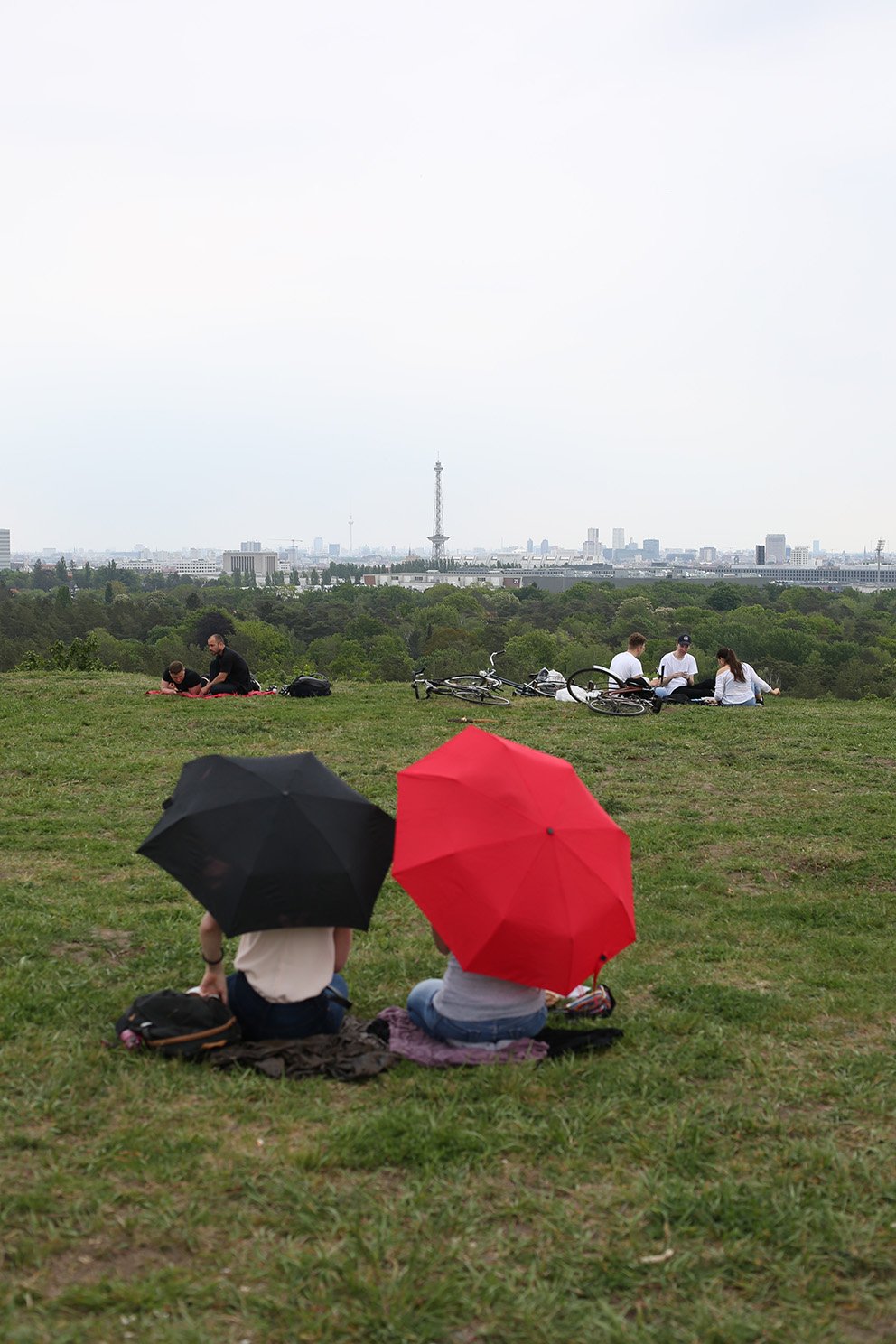 Several people sit on a hill with umbrellas, looking out over the distant city skyline with forest in the foreground.