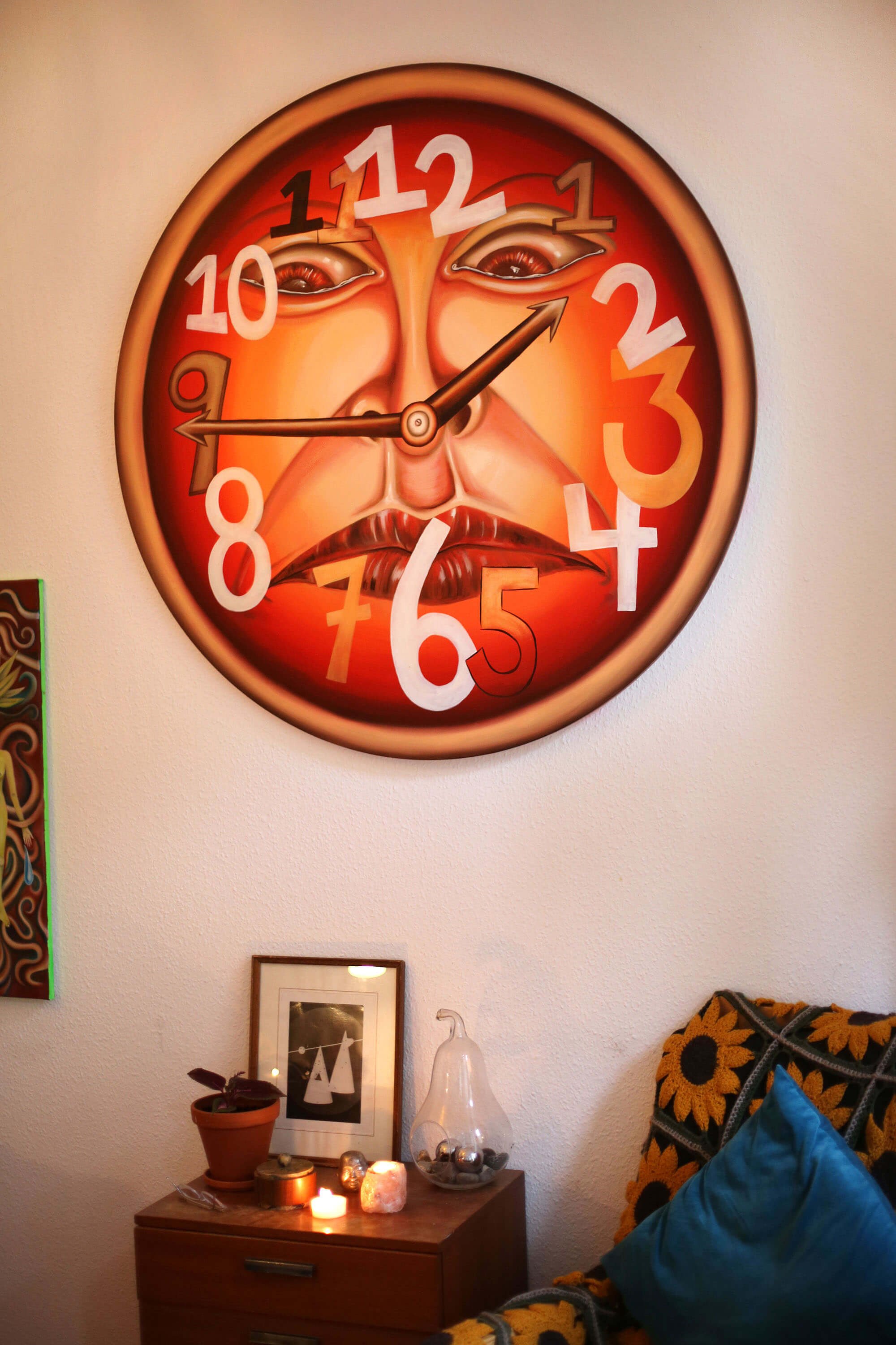Round wall artwork shaped like a clock, featuring an expressive face on the dial; below it a small sideboard with candles, plants, and decorative objects.