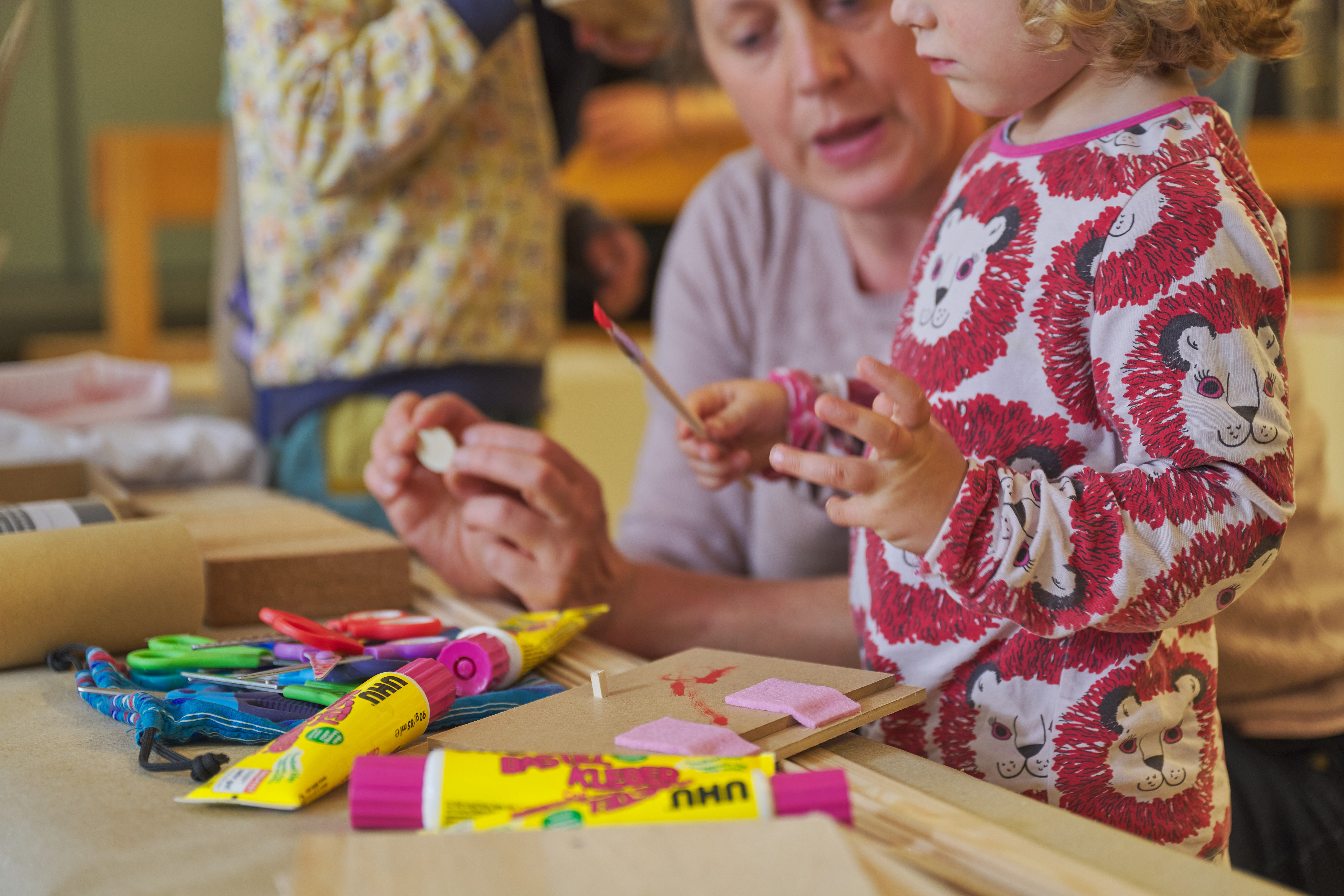 Several children are working together on a craft project at a table. One child cuts with scissors while another presses a pencil onto a piece of paper. Cardboard, tools, and craft materials are spread across the table. The scene is captured in warm lighting.