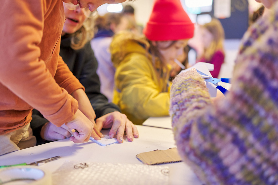 Several children are working together on a craft project at a table. One child cuts with scissors while another presses a pencil onto a piece of paper. Cardboard, tools, and craft materials are spread across the table. The scene is captured in warm lighting.
