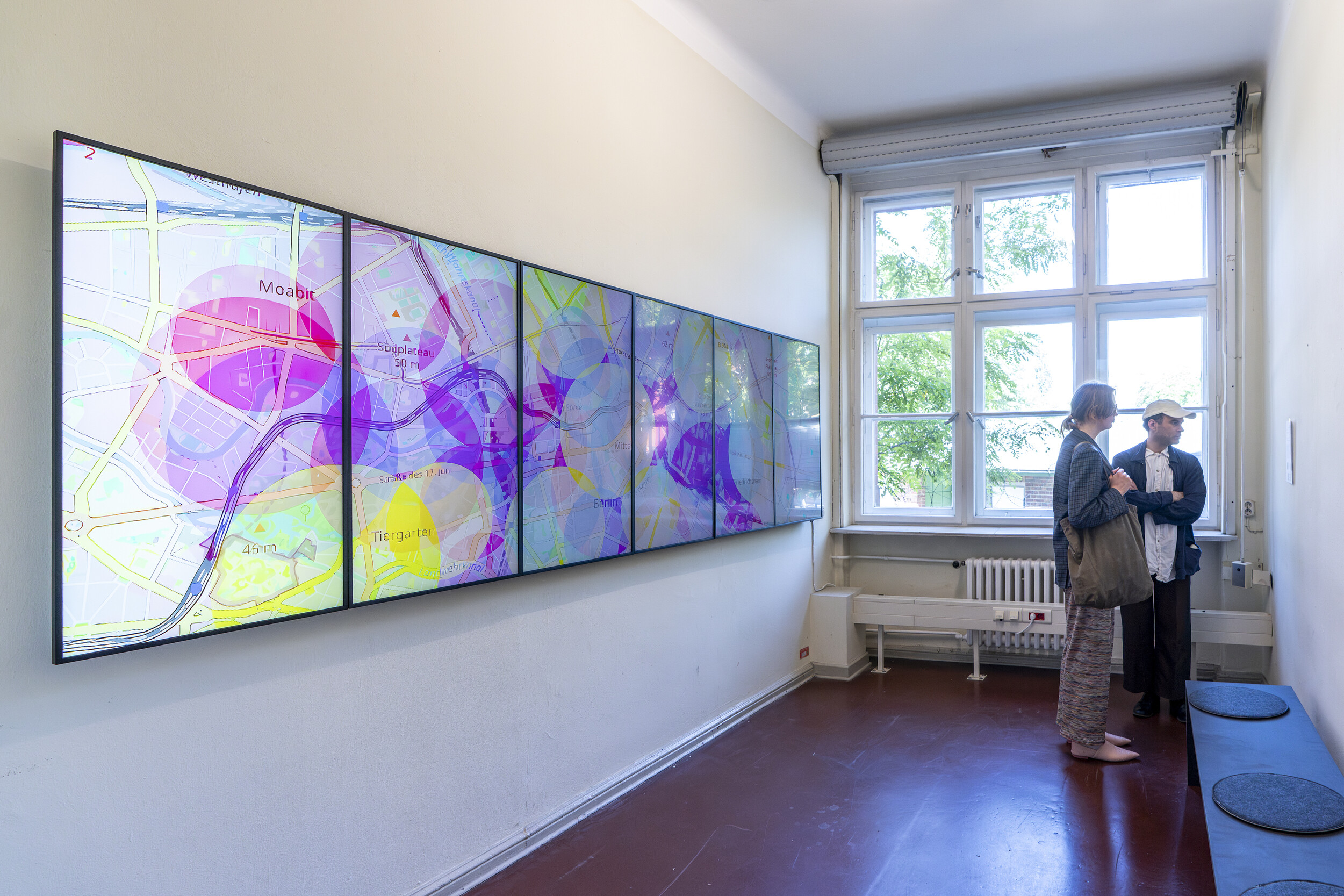 Narrow exhibition space with a large multi-screen installation mounted on the left wall. The adjacent displays show a vividly colored cartographic image with overlapping circles and street networks. Two visitors stand talking near the window on the right side. Daylight fills the room, and a long bench runs along the right wall.