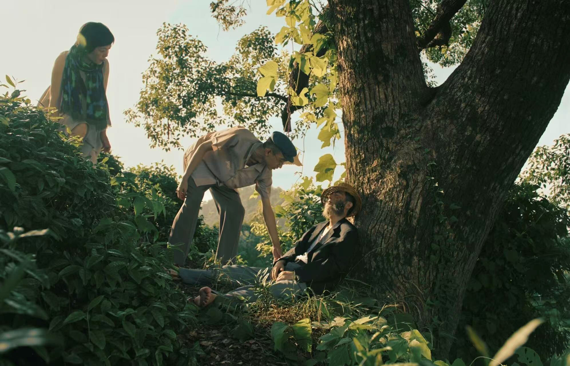 An older man is leaning against a thick tree trunk, apparently asleep. A younger man is leaning down toward him and touching his arm; a woman is standing nearby, watching the scene.