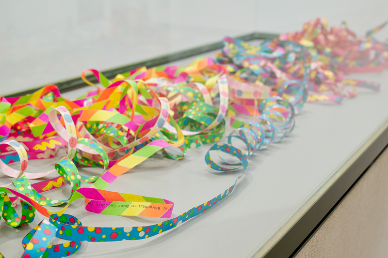 Auf einem Tisch liegen zahlreiche bunte Papier- und Kunststoffstreifen mit Punkten und Mustern, die zu lockeren Schlaufen und Spiralen geformt sind. Die farbenfrohen Bänder verteilen sich über die gesamte Oberfläche.
