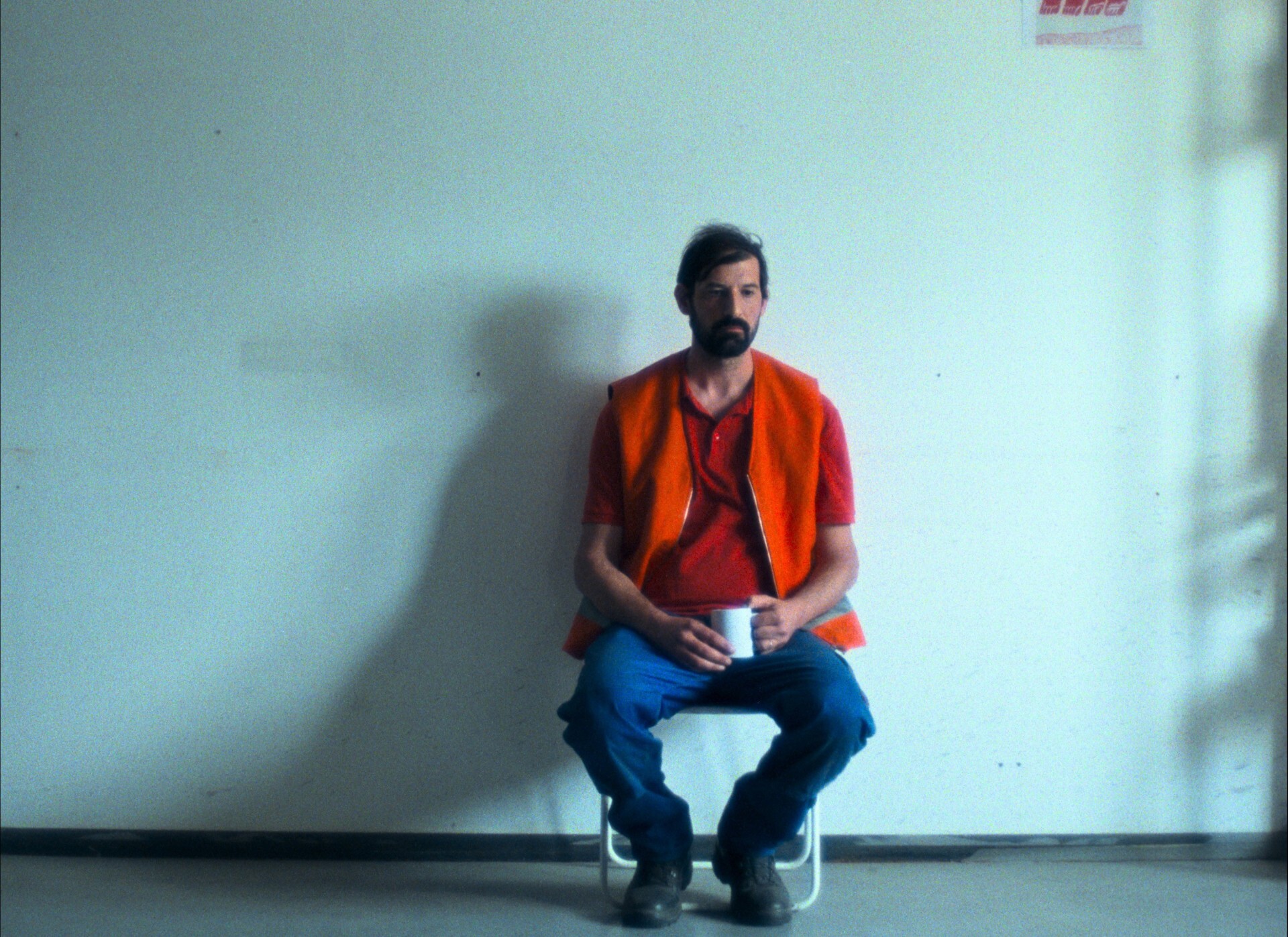 Eine Person sitzt frontal auf einem einfachen Stuhl vor einer leeren weißen Wand und hält eine weiße Tasse in den Händen. Sie trägt ein rotes Shirt, eine orangefarbene Warnweste und blaue Arbeitshosen. Das Bild wirkt leicht unscharf und körnig, mit weichem Licht und einem langen Schatten an der Wand, wodurch eine stille, nachdenkliche Atmosphäre entsteht.