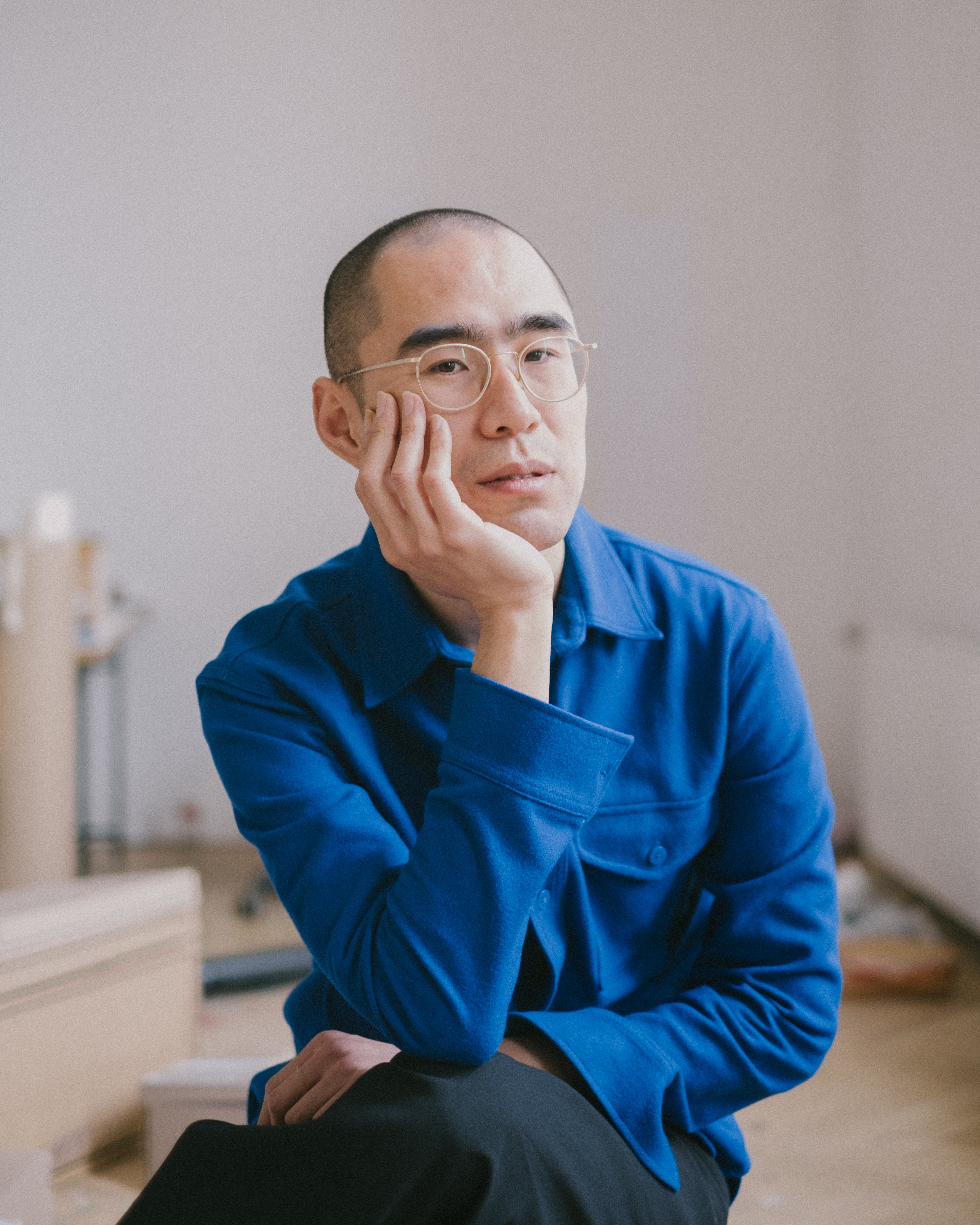 Ein Mann mit strahlend blauem Hemd stützt seinen Kopf im Sitzen auf seine Hand. Er blickt in die Kamera, trägt eine Brille und hat sehr kurze schwarze Haare.