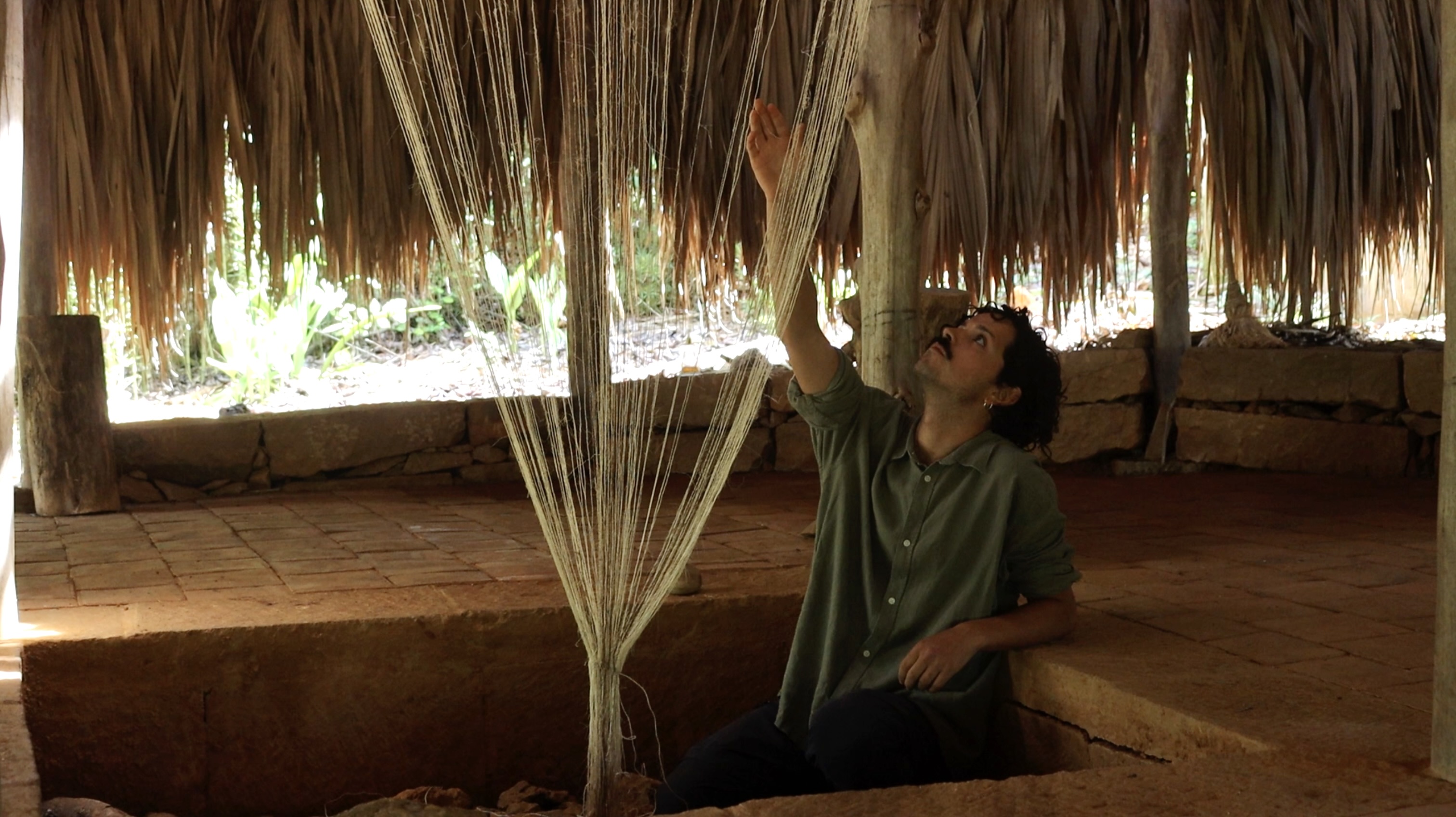 Ein Mann mit dunklen Haaren und einem Schnurrbart sitzt einer Grube unter einem Strohdach und berührt hängende Fasern.