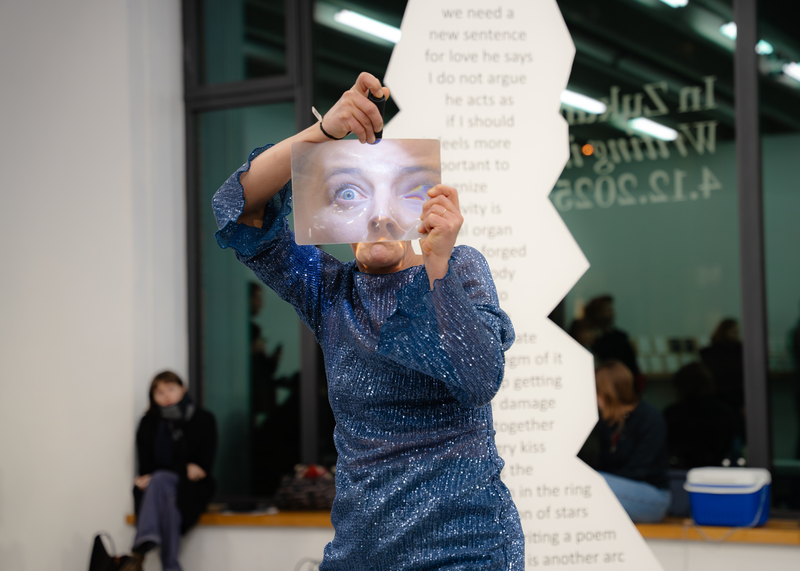 Eine performende Person in einem glitzernden blauen Kleid hält ein Blatt mit dem vergrößerten Bild eines Gesichts vor ihr eigenes Gesicht. Im Hintergrund sind Zuschauer*innen sowie ein großer, unregelmäßig geformter Textträger mit englischem Schriftzug zu sehen.