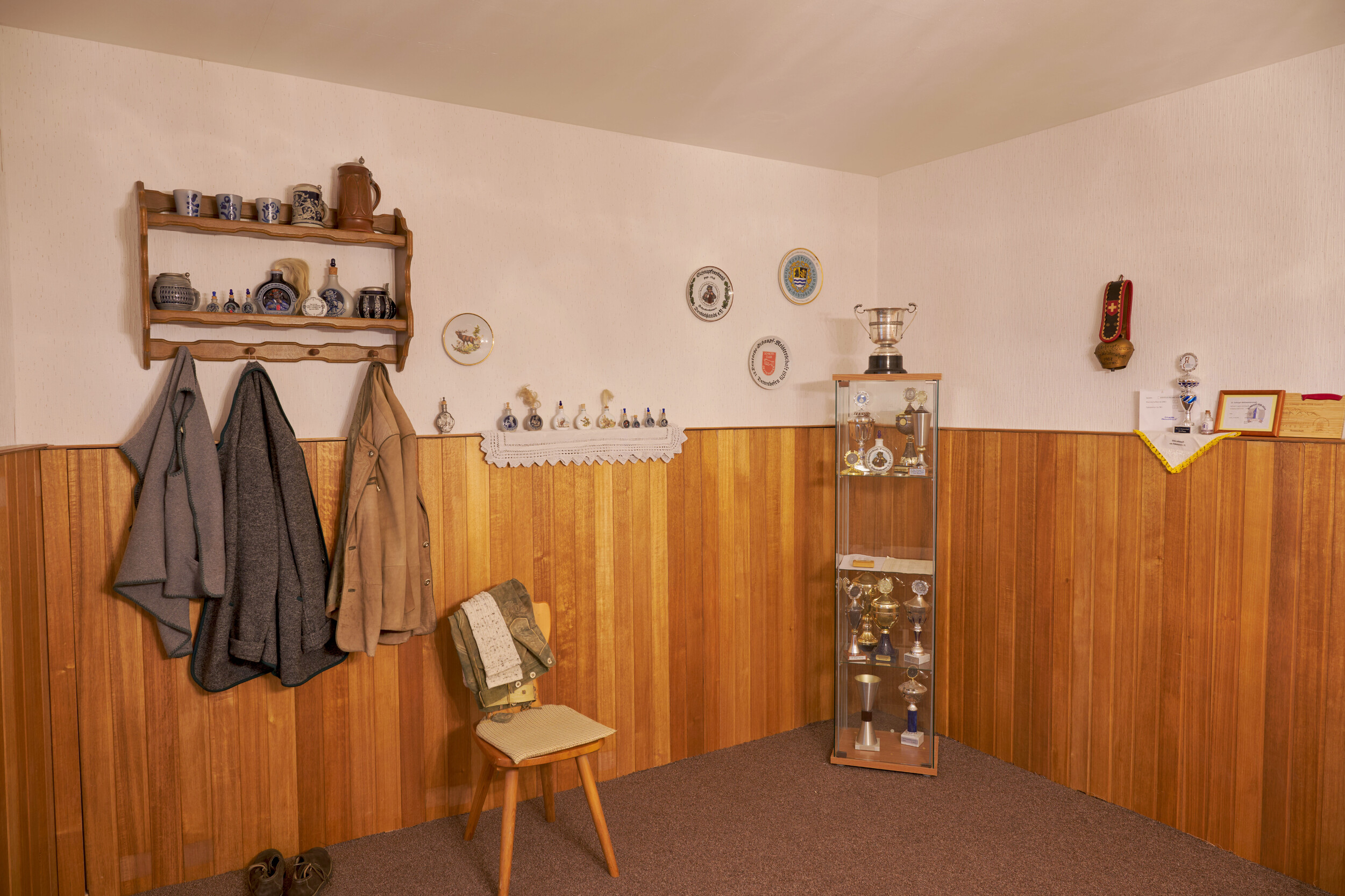 Ecke eines Raums mit Holzvertäfelung, einem Stuhl, einer Garderobe mit Jacktes und einer Vitrine mit Pokalen.