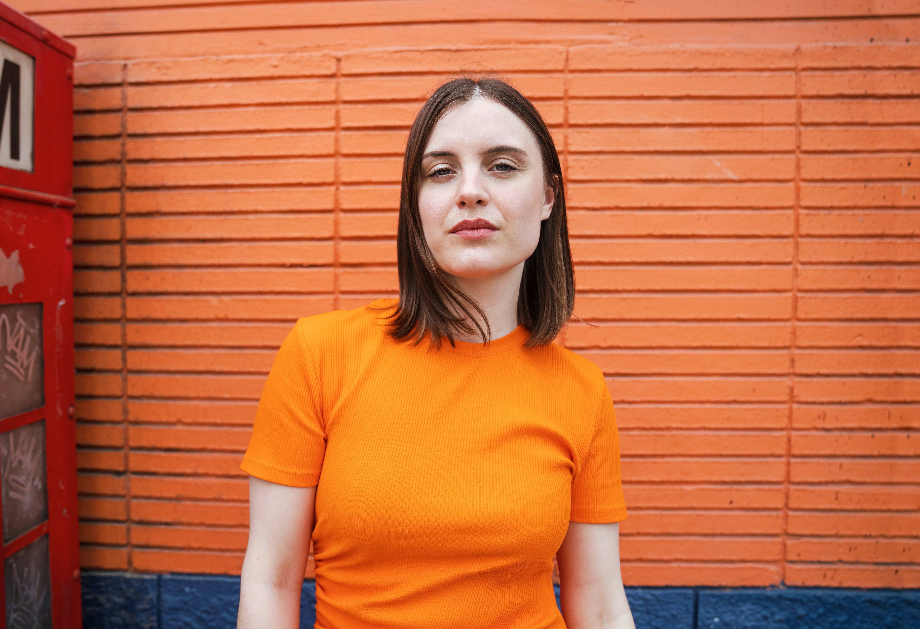 Eine Frau mit orangefarbenem T-Shirt und dunklen, schulterlangen Haaren blickt in die Kamera. Ihr Kopf ist leicht nach oben geneigt und sie steht vor einer ebenfalls orangefarbenen Wand.