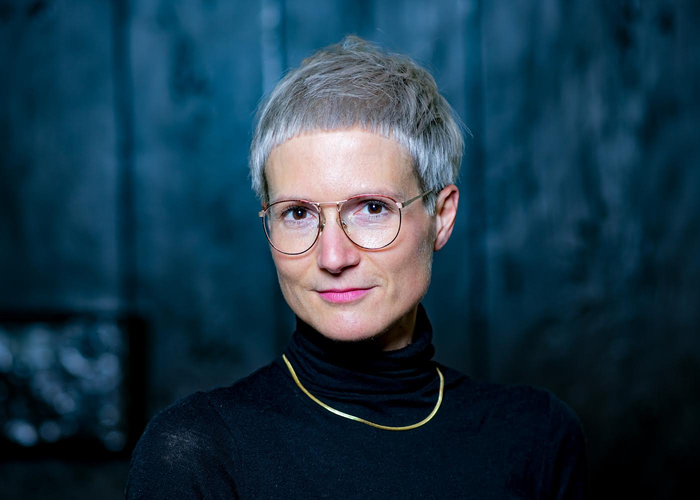 Eine Frau mit kurzen weißen Haaren lächelt leicht in die Kamera. Sie trägt eine Brille mit rundem, dünnen Metallgestell, eine schlichte Goldkette und ein schwarzes Rollkragenoberteil.