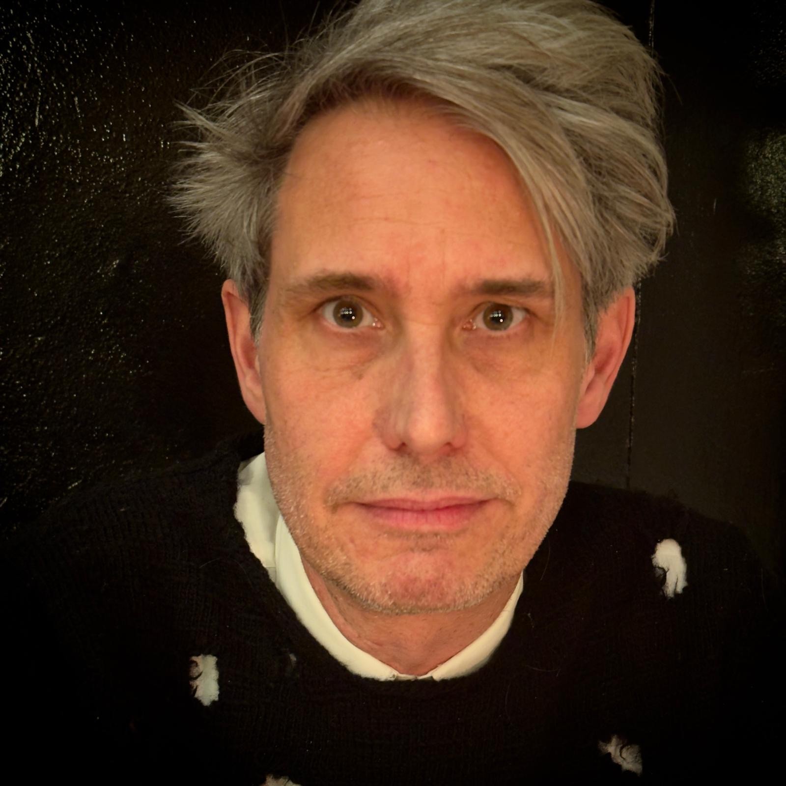 Ein Mann mit Dreitagebart blickt in die Kamera. Er hat kurze, vorne ins Gesicht fallende Haare und trägt einen schwarzen Pullover mit weißen Sprenkeln darauf.