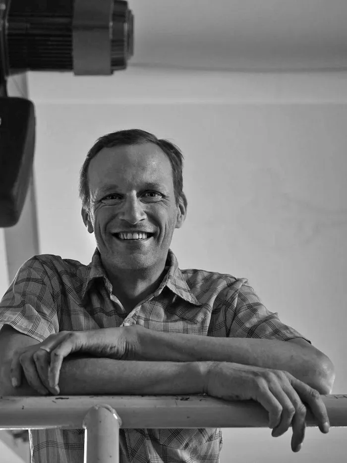 Black-and-white photograph of a smiling person in a checkered shirt leaning on a railing. Chains hang from a technical device in the background, and the space appears industrial.