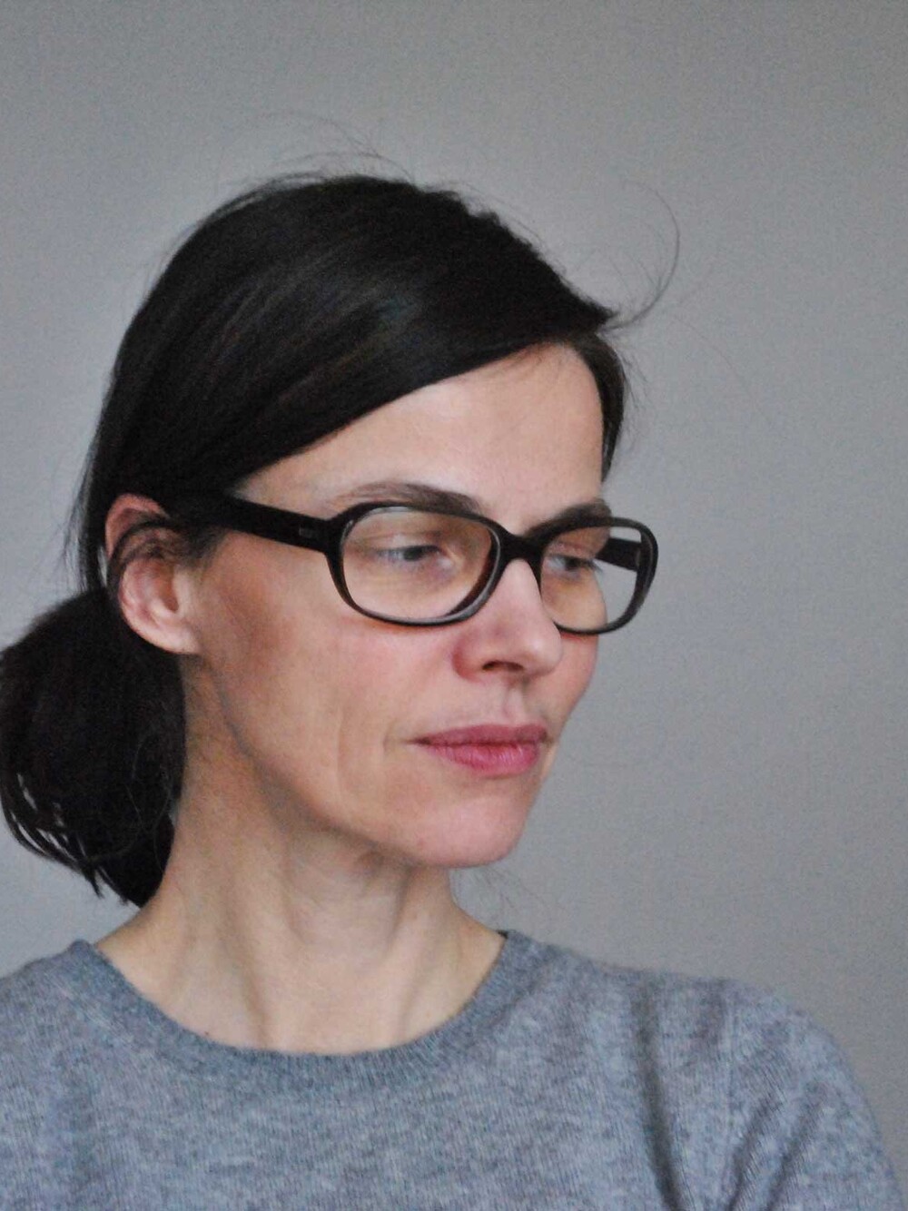 Portrait of a person with dark hair and black glasses, looking downward to the side. The person is wearing a gray sweater, and the background is a plain gray color.