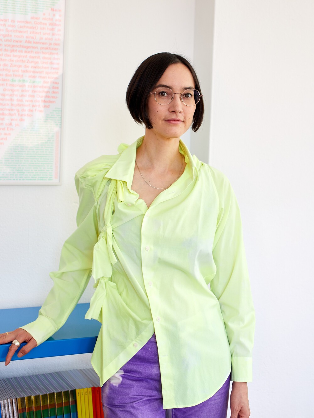 Eine Frau mit kurzen dunklen Haaren und einer Brille mit dünnem Metallgestell steht an einem niedrigen Bücherregal. Sie trägt eine hellgelbe Bluse und eine lilafarbene Hose.