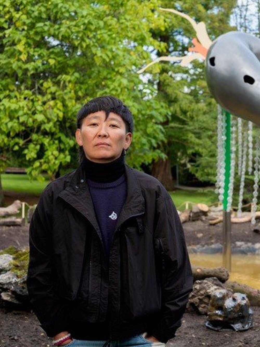 Eine Person steht im Freien vor einer großen, farbigen Skulptur mit hängenden Kettenelementen und organischen Formen. Im Hintergrund sind Bäume und ein Gartenbereich zu sehen.