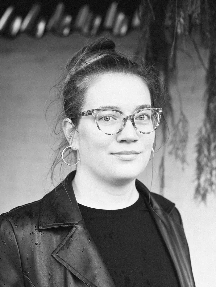 Schwarz-weißes Porträt einer Person mit heller Brille, hochgestecktem Haar und dunklem Oberteil. Die Person blickt leicht lächelnd nach vorne; im Hintergrund sind unscharfe Pflanzen zu sehen.