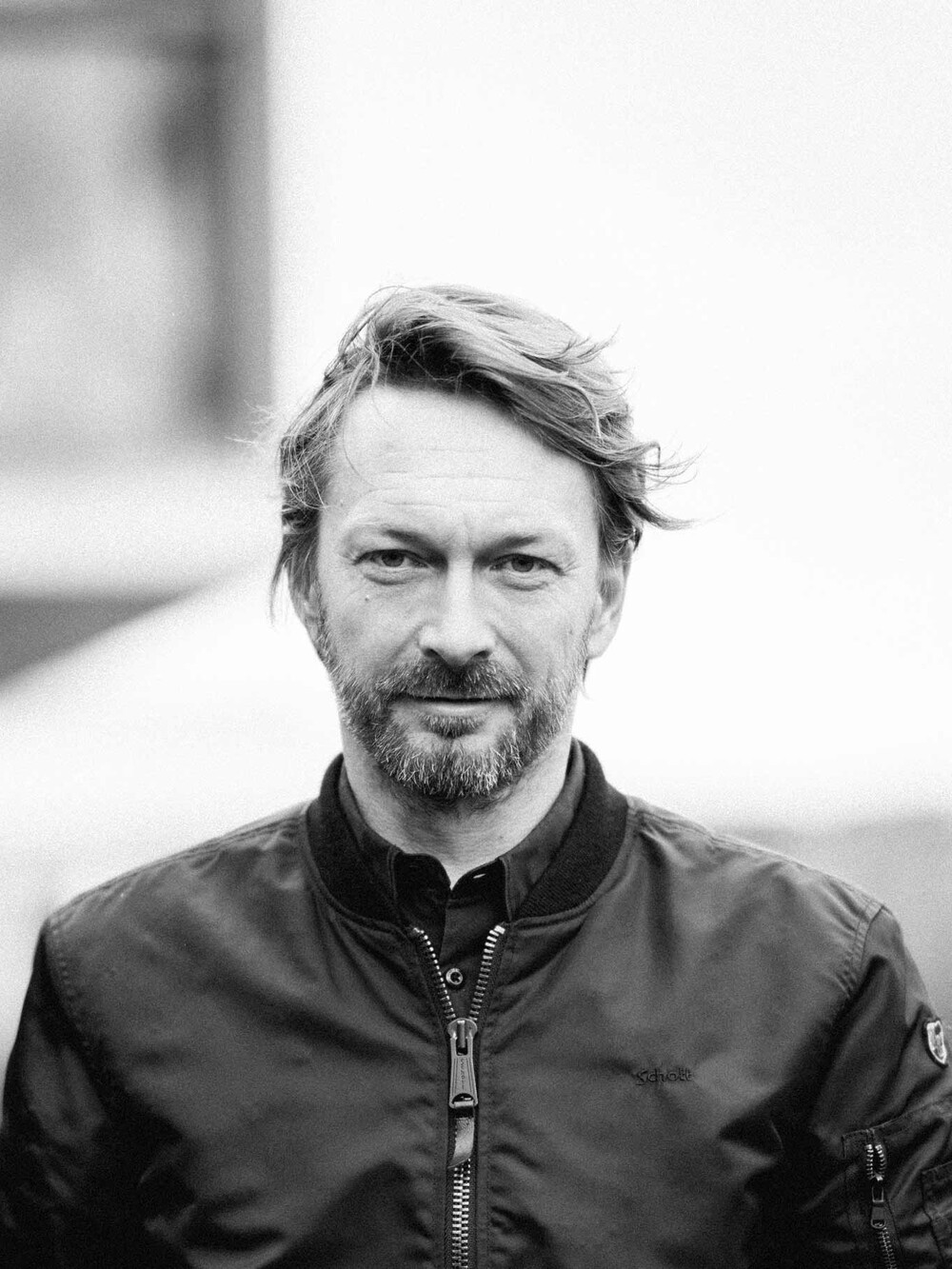 Schwarz-weißes Porträt einer Person im Freien, die in die Kamera blickt. Die Person trägt eine dunkle Jacke, der Hintergrund ist unscharf und zeigt eine Außenlandschaft.