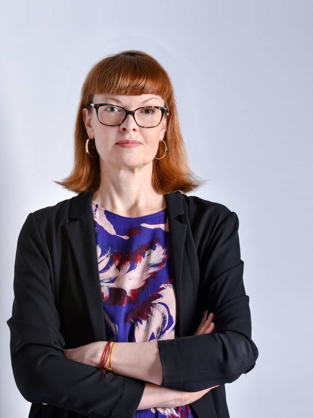 Portrait of a person with shoulder-length reddish hair and glasses, standing with arms crossed against a light background. The person is wearing a dark jacket and a patterned top.