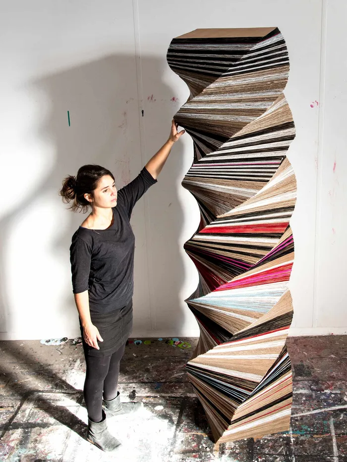 Artist standing in a studio next to a tall, twisted sculpture made of layered cardboard or paper materials.