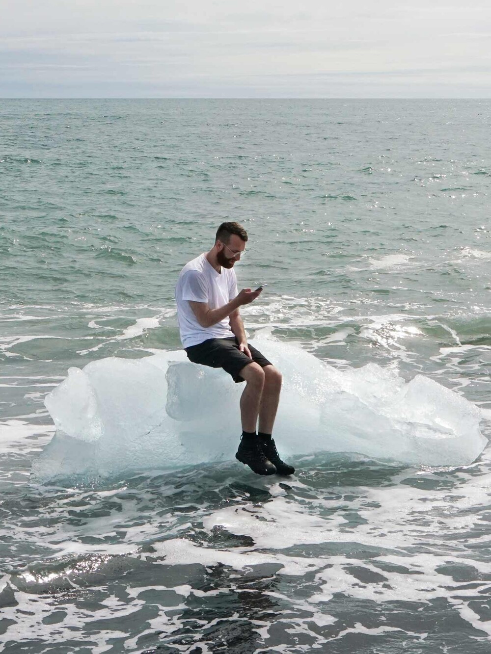 Ein Mann sitzt auf einem Eisblock im Meer und blickt auf sein Smartphone, während Wellen um ihn herum an den Strand schlagen.