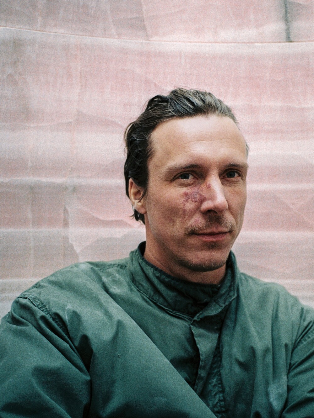 Nahporträt eines Mannes mit zurückgekämmtem Haar und ernster Miene vor hellem, strukturiertem Hintergrund.