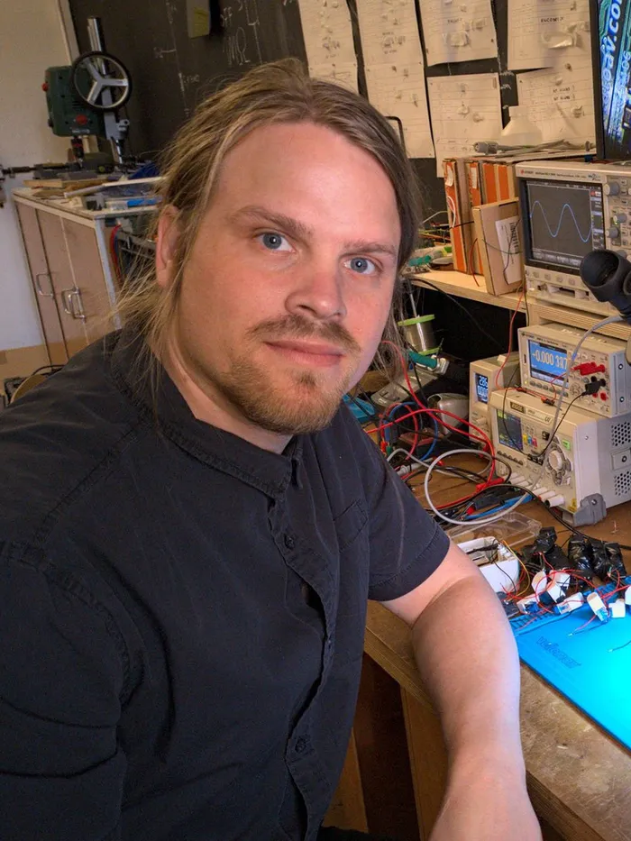 Man with shoulder-length hair seated at an electronics workbench with measuring devices and a microscope, a circuit board illuminated in front of him.