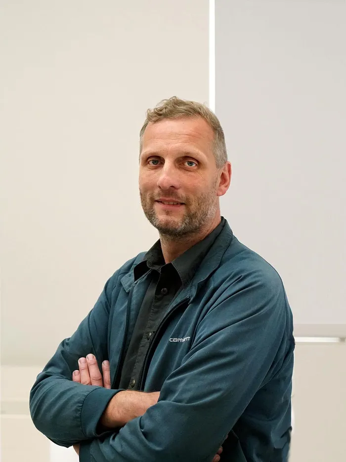 Mann mit kurzem Haar und Bart steht mit verschränkten Armen vor hellen Fenstern in einem modernen Innenraum.