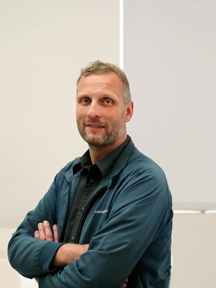 Man with short hair and a beard stands with arms crossed in front of bright windows in a modern interior.