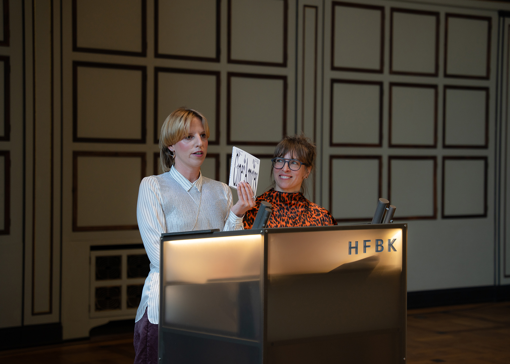 Zwei Frauen stehen hinter einem beleuchteten Rednerpult mit der Aufschrift „HFBK“ in einem Saal mit dekorativen Wandpaneelen. Eine der Frauen hält ein Buch hoch und spricht, die andere lächelt. Auf dem Pult stehen Mikrofone. Der Raum wirkt festlich und gut ausgeleuchtet.