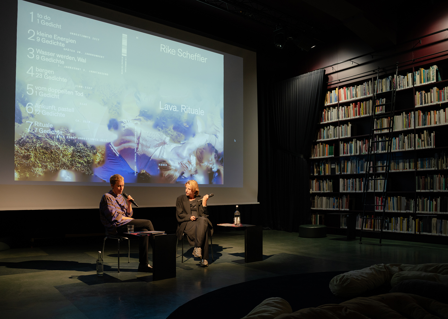 Zwei Personen sitzen auf einer Bühne in einem Bibliotheksraum und führen ein Gespräch. Beide halten Mikrofone in der Hand. Hinter ihnen ist eine große Projektion mit dem Titel „Rike Scheffler – Lava. Rituale“ sowie einer Liste von Gedichten zu sehen. Rechts im Raum stehen hohe Bücherregale mit einer Leiter, das Licht ist gedämpft und konzentriert sich auf die Bühne.