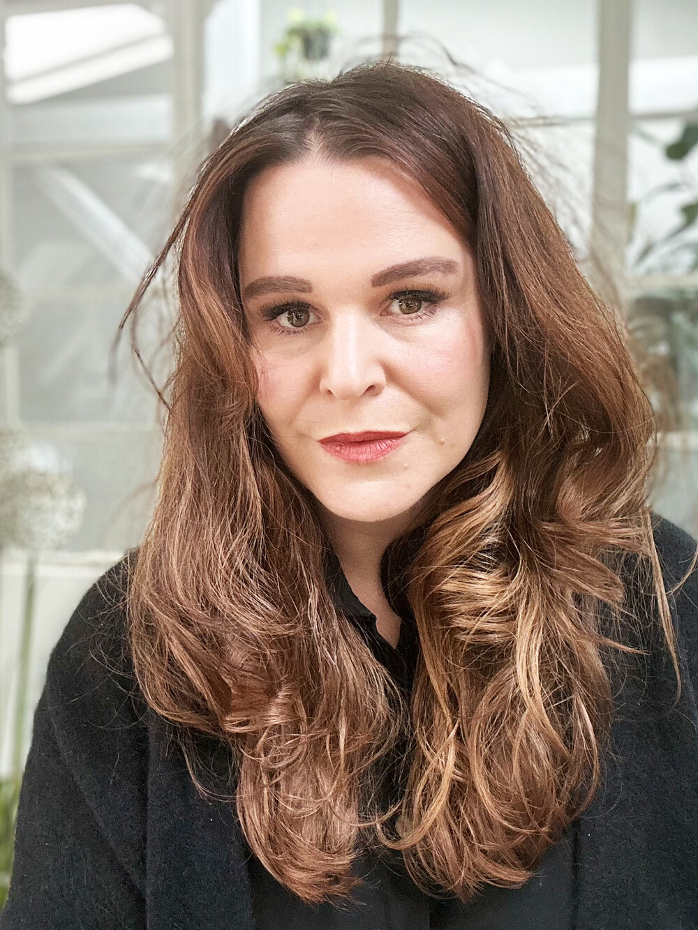 Portrait of a woman with long, wavy hair looking at the camera in a bright interior with plants in the background.