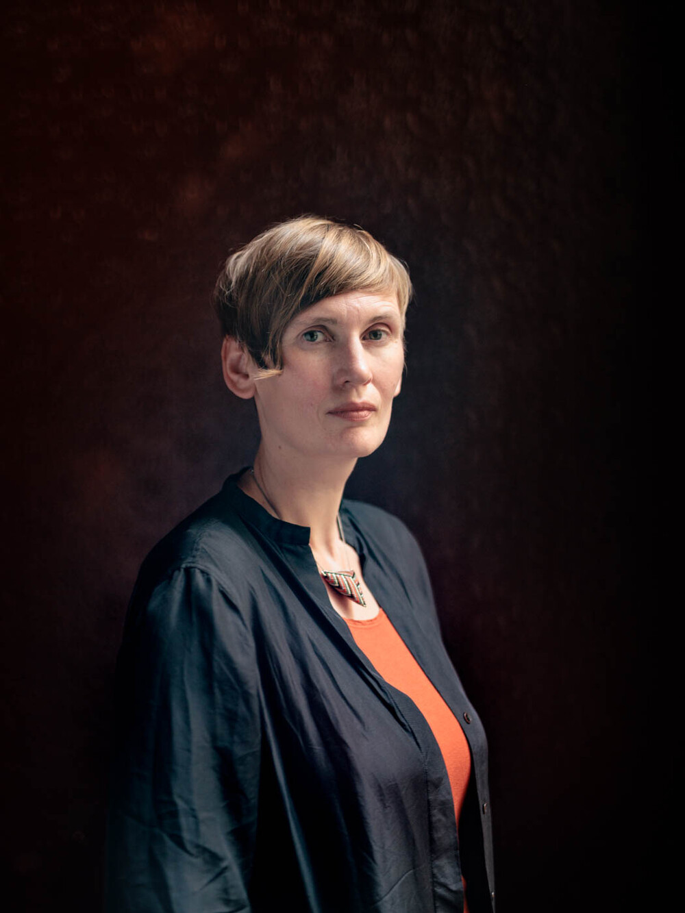 Eine Frau mit kurzen dunkelblonden Haaren blickt in die Kamera, sie trägt eine Kette und eine schwarze Bluse mit orange-farbenem Oberteil darunter.