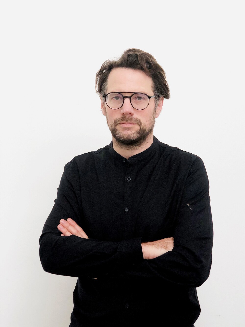 Porträt eines Mannes mit Brille und Bart, der mit verschränkten Armen vor einem weißen Hintergrund steht.