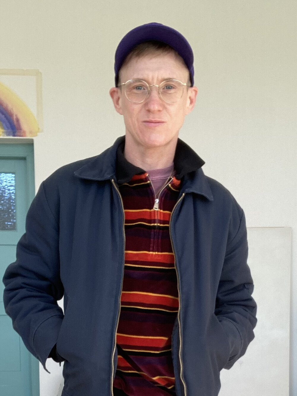 Ein Mann mit blauer Cappy, einer Brille mit durchsichtigem Gestell und kurzen blonden Haaren blickt mit einem leichten Lächeln in die Kamera. Er trägt ein rot-orange gestreiftes Oberteil und darüber eine dunkelblaue Jacke.