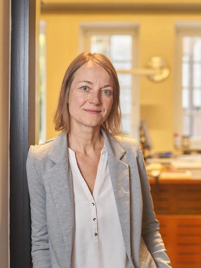 Porträt einer Frau in hellem Blazer und weißer Bluse, die in einer geöffneten Tür zu einem Büro steht und in die Kamera blickt.