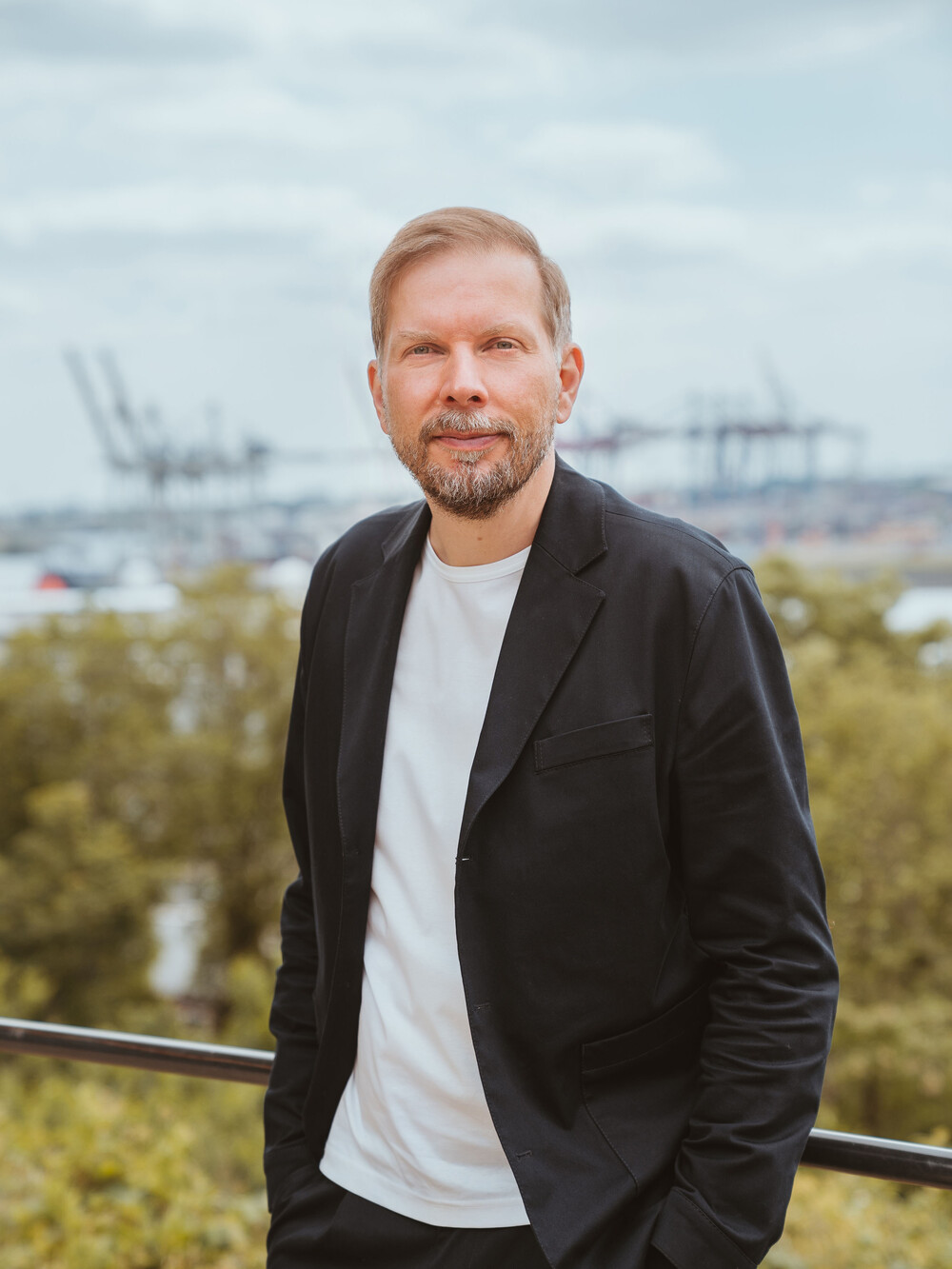 Ein Mann mit kurzem blondem Haar und Bart steht draußen auf einer Terrasse. Er trägt ein schwarzes Sakko über einem weißen T-Shirt und hat die Hände in den Hosentaschen. Im Hintergrund sind Bäume, Wasser und Hafenkräne zu sehen.