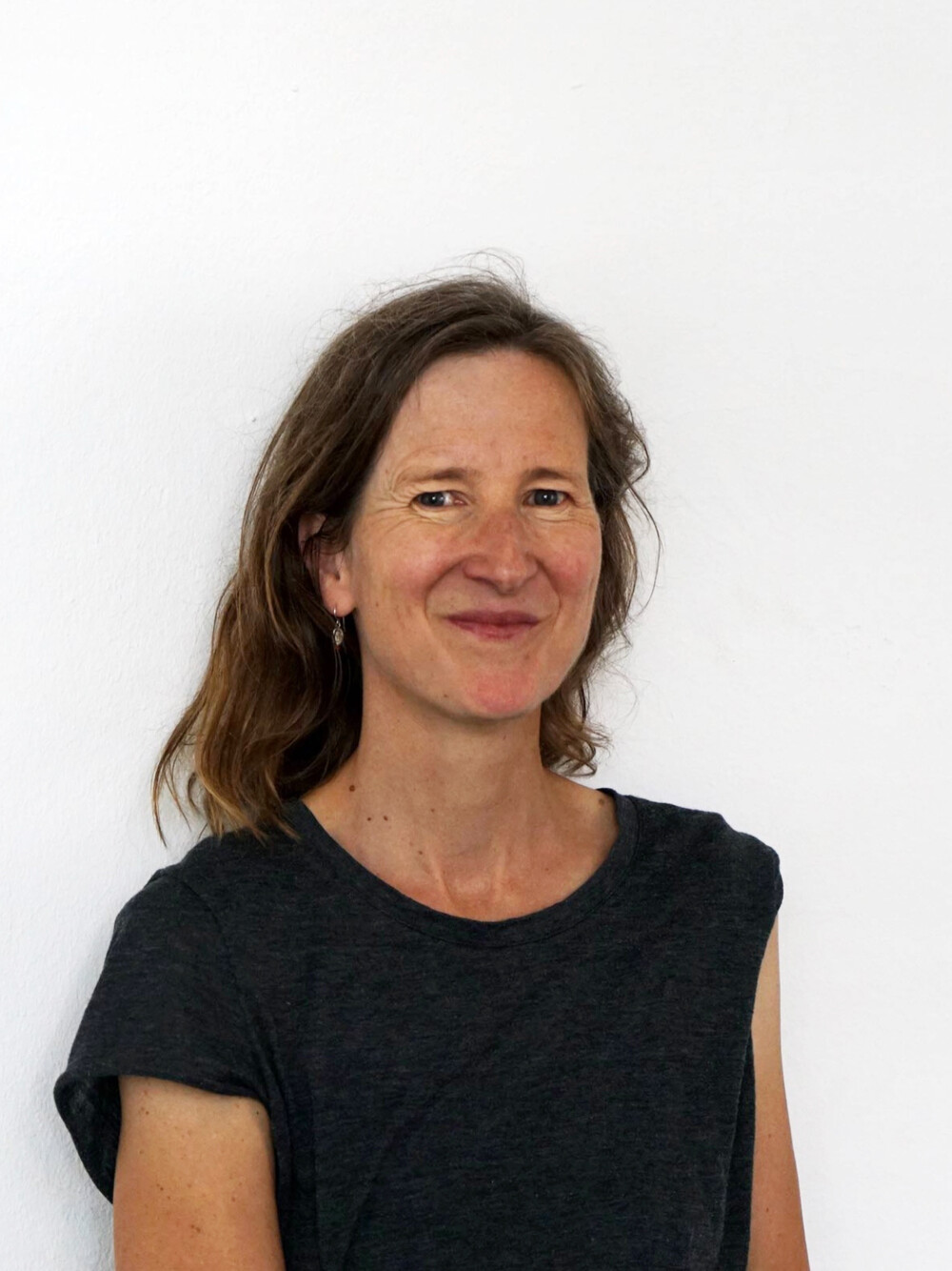 Porträt einer Frau mit schulterlangem Haar, lächelnd, vor weißem Hintergrund, trägt ein ärmelloses Oberteil.