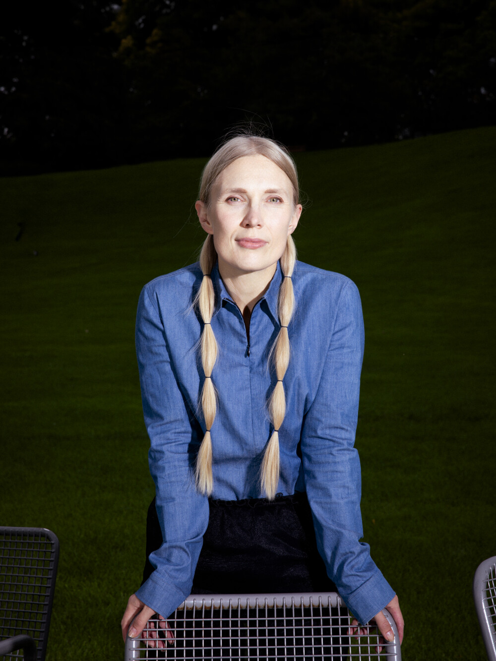 Eine Frau mit langen blonden Zöpfen und einem blauen Hemd stützt sich mit beiden Händen auf die Metalllehne eines Stuhls.