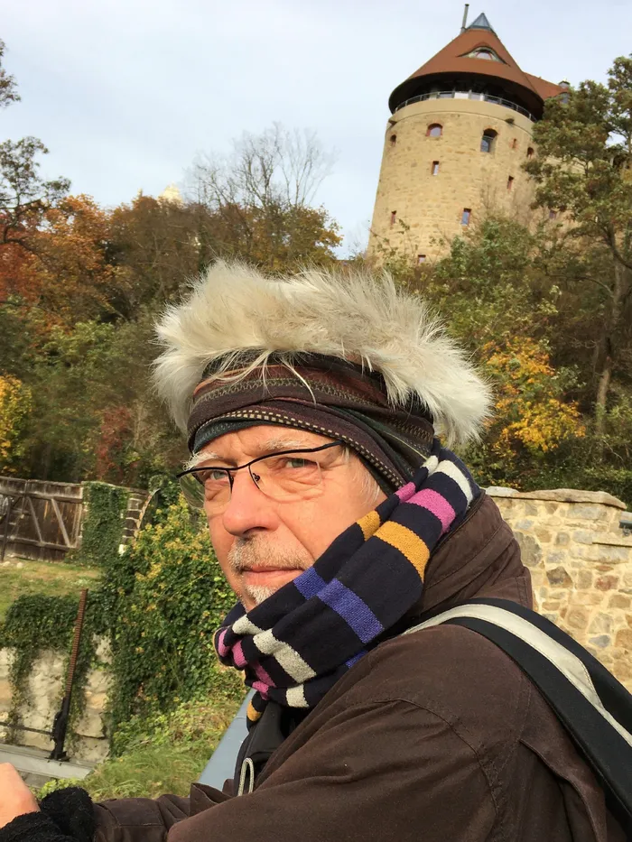 Portrait of a man wearing glasses, a scarf, and a headband outdoors, with a round stone tower and autumn trees in the background.