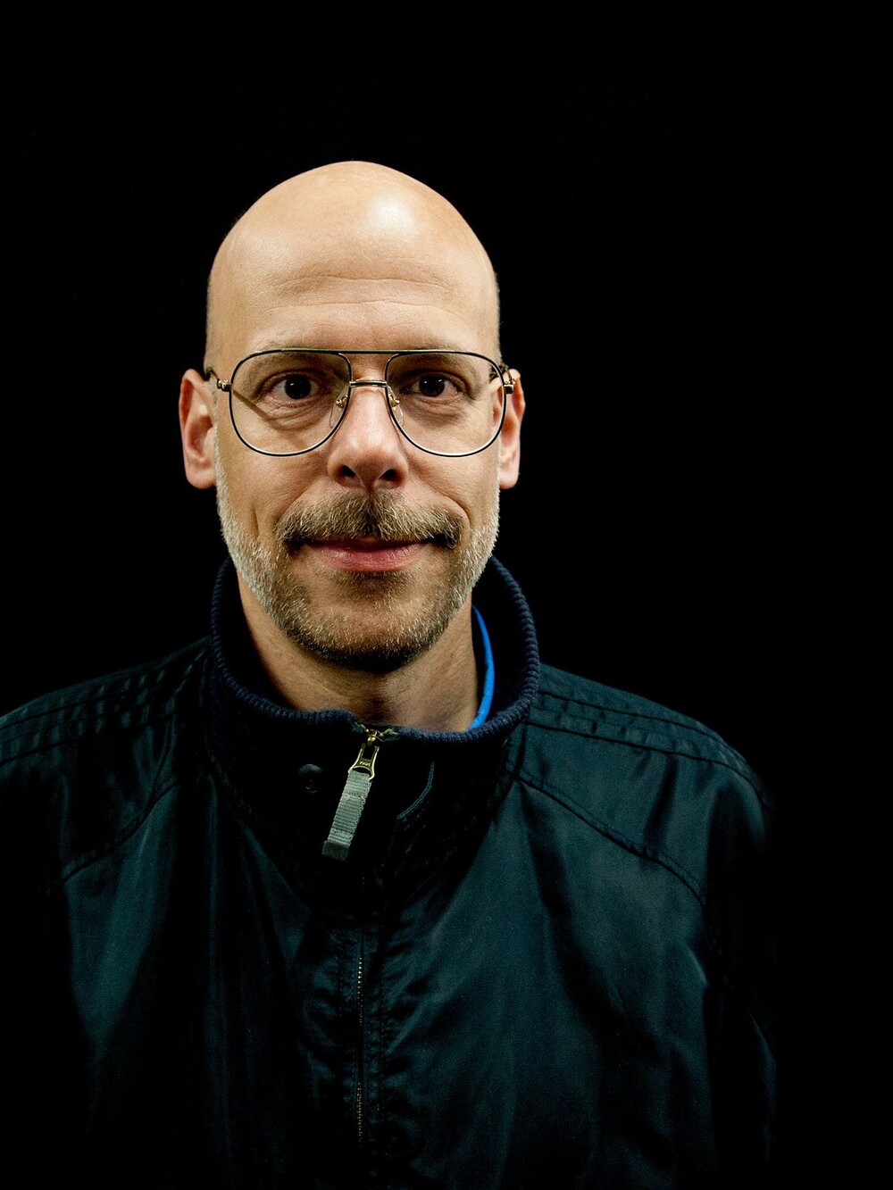 Portrait of a bald man wearing glasses, photographed frontally against a black background.