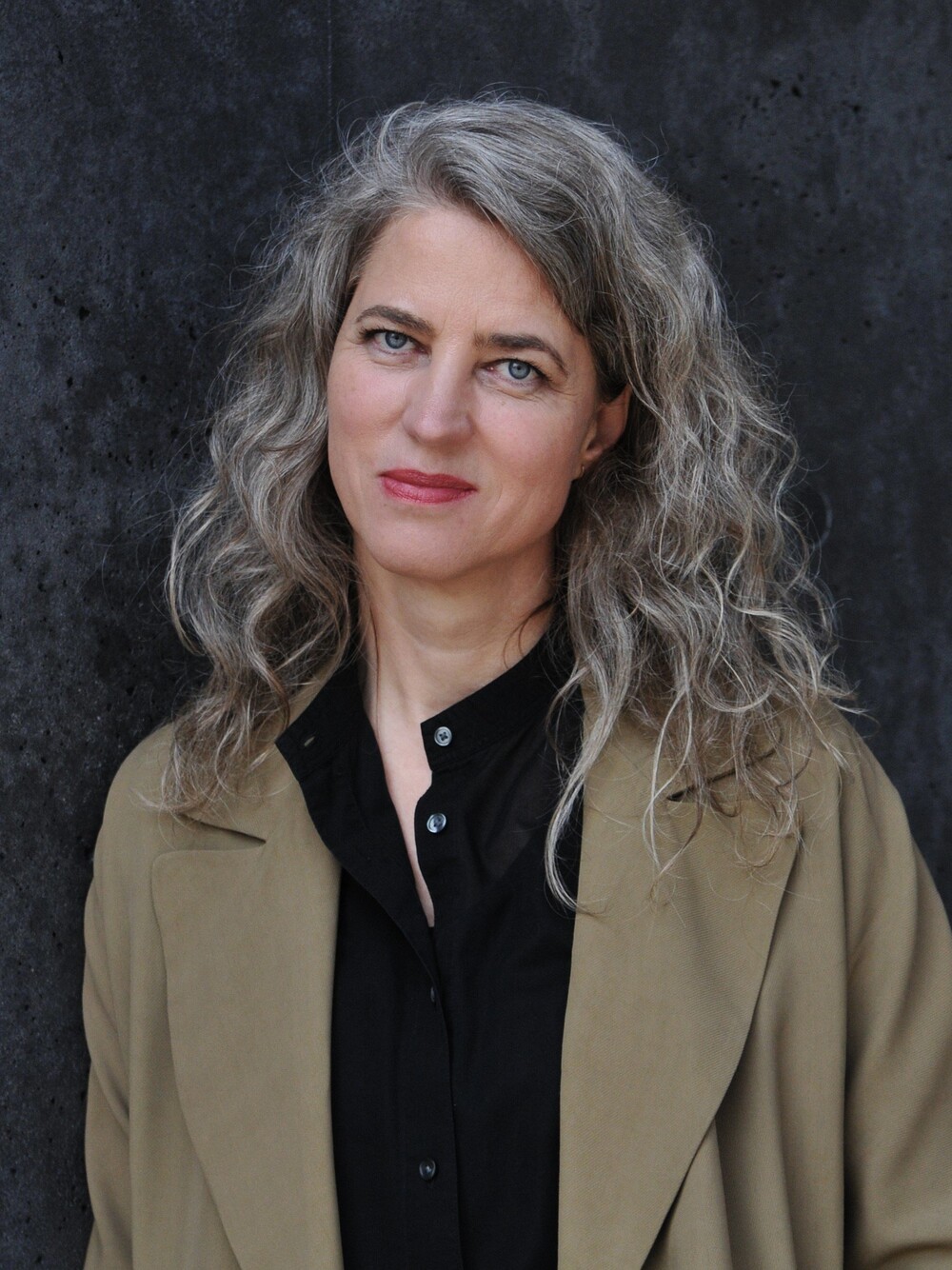 Porträt einer Frau mit grauem, welligem Haar vor dunkler Betonwand, leicht zur Seite gedreht.
