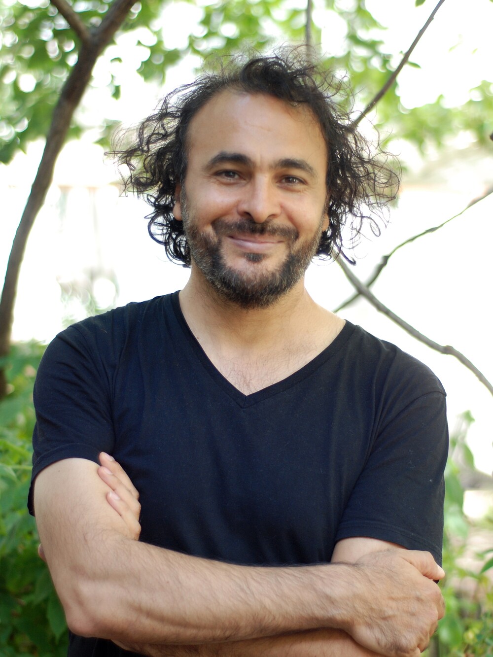 Porträt eines lächelnden Mannes mit lockigem Haar und Bart, die Arme verschränkt, im Freien vor Bäumen.